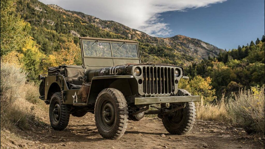 A restored classic army surplus jeep parked on a dirt trail in a natural U.S. landscape. Olive drab paint, fold-down windshield, exposed spare tire, and rugged tires clearly visible.
