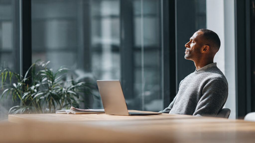 A clean, text-free image of a working professional at a desk practicing calm breathing with a laptop nearby, natural light, modern office setting, relaxed posture.