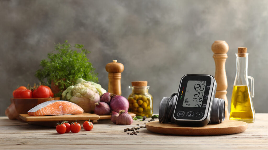A clean, text-free image showing a home blood pressure monitor on a table beside heart-healthy foods like vegetables, fish, and olive oil. Natural lighting, realistic style.